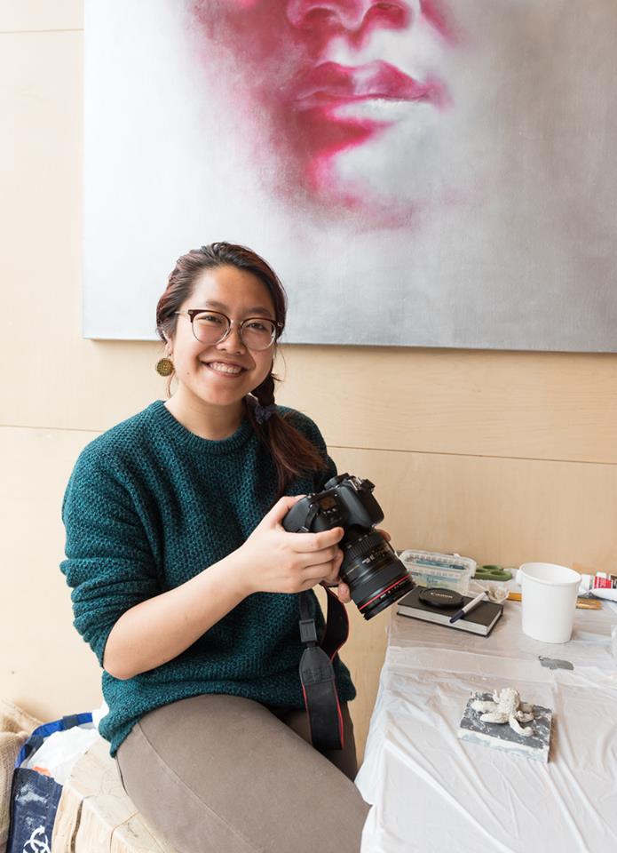 Christie Wong sits on a stall by a table covered in cloth, holding a DSLR camera and smiling at the lens of the camera taking the photo. She is an Asian women wearing round, wooden earrings, round glasses, a green sweatshirt and grey/beige pants.