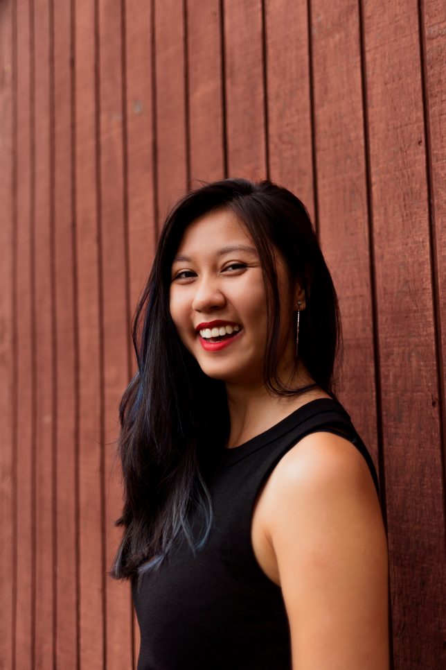 Linh, an Asian woman with chest-length black hair, is looking at the camera from the side. She stands by a wooden-paneled wall and is smiling with her mouth open. She's wearing a black, sleeveless shirt and red lipstick.